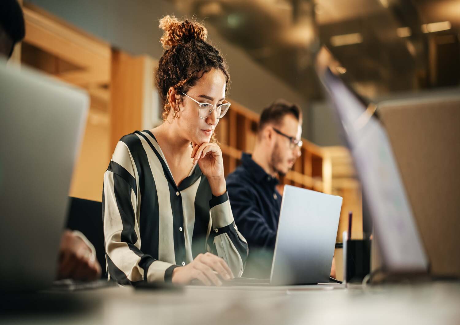 Woman wearing glasses and looking focused at her laptop screen while working at an office with her colleagues.