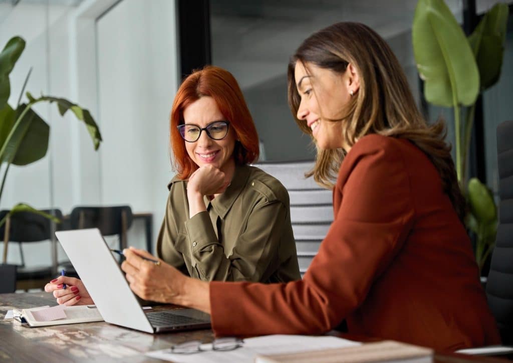 Two female coworkers sitting down in front of a laptop while one of them trains the other on a specific process.